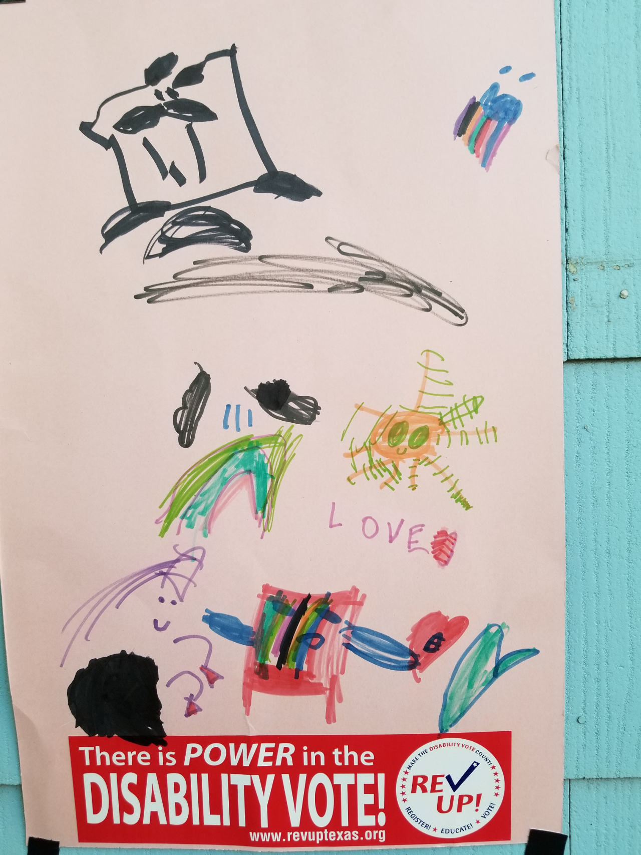 Sign with kid’s drawing of wheelchairs, love and symbols with a Rev Up Disability Vote bumper sticker on the bottom of the sign
