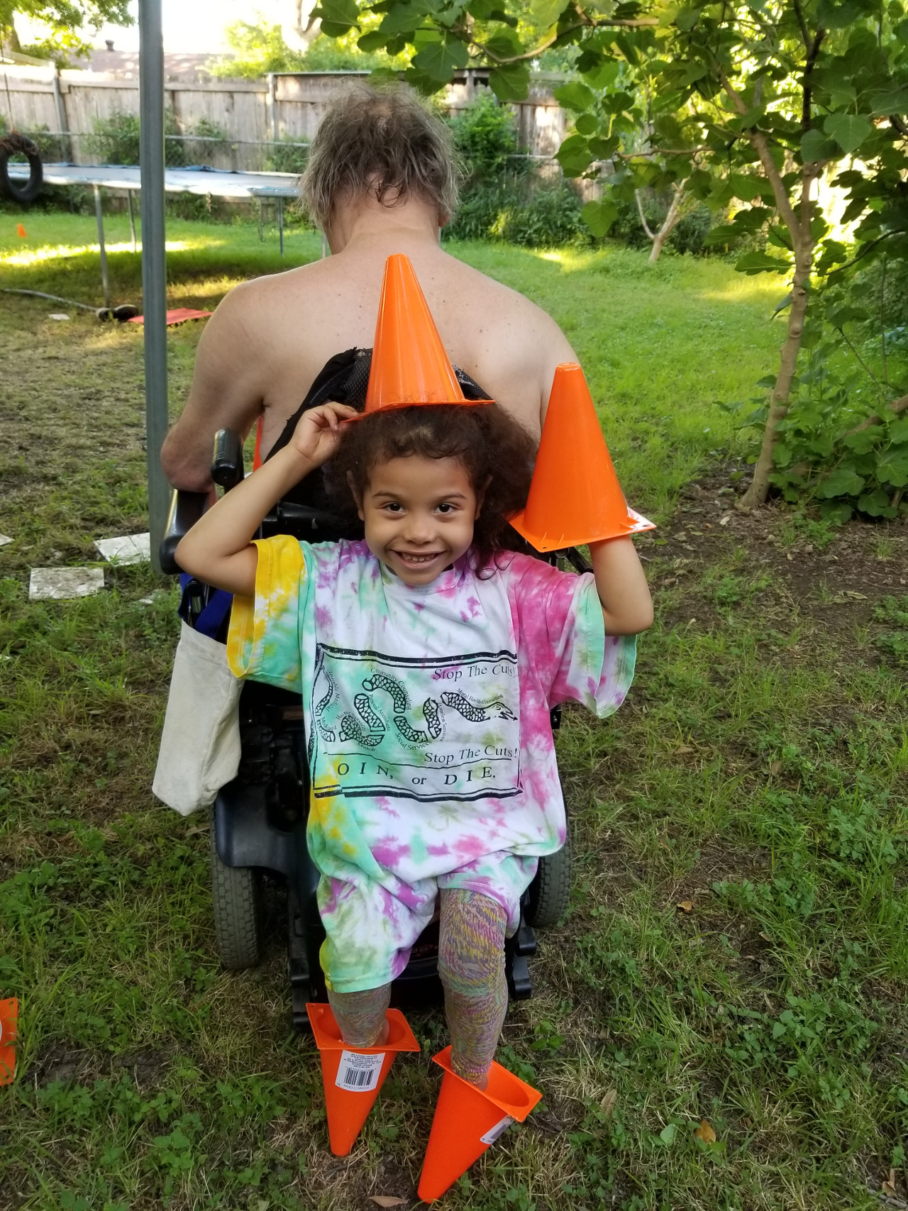 Young girl sits on the back of her grandpa’s motorized wheelchair. She is smiling and has little traffic cones on her head, feet and hand.